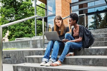 Two young female college students sitting on steps in front of a university building, looking at a laptop screen together.